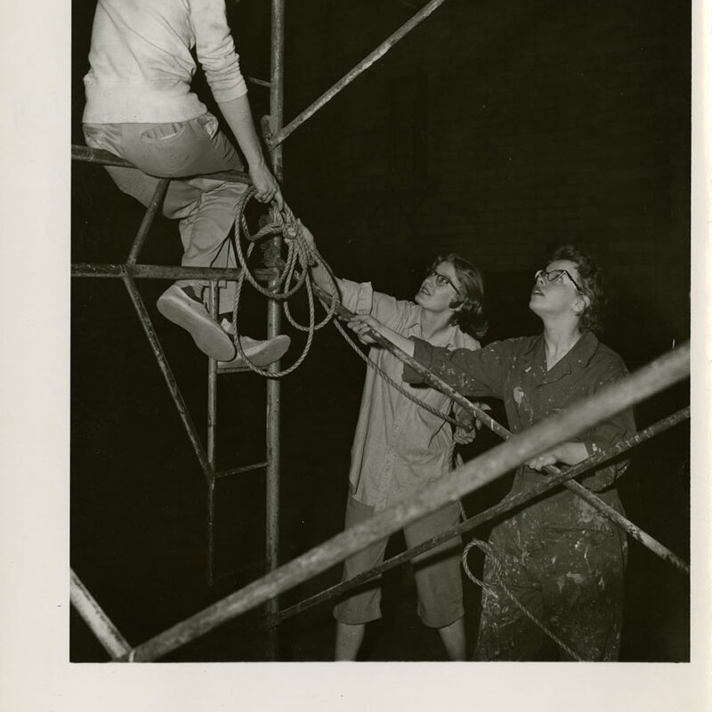 Two students hand rope to student sitting on scaffolding